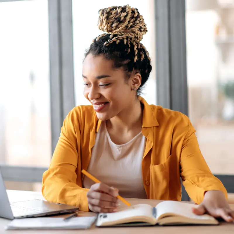 Educación online, e-learning. Joven afroamericana feliz con ropa informal y elegante, estudiando a distancia, usando un portátil, escuchando clases online, tomando apuntes mientras está en casa, sonríe